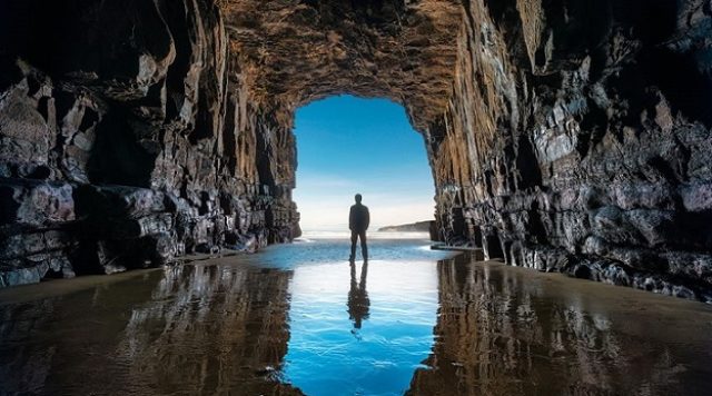 A picture from within a dark cave, showing a man standing at the entrance