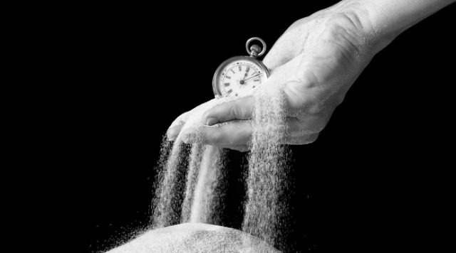A hand holding a small clock with sand slipping through the fingers into a pile of sand below