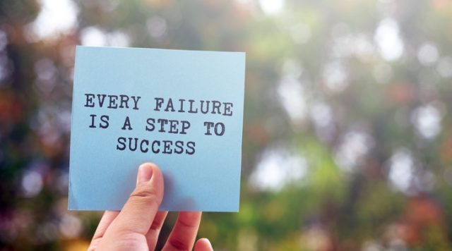 A person's hand holding a sticky note that says "Every failure is a step to success", set against an outdoor background