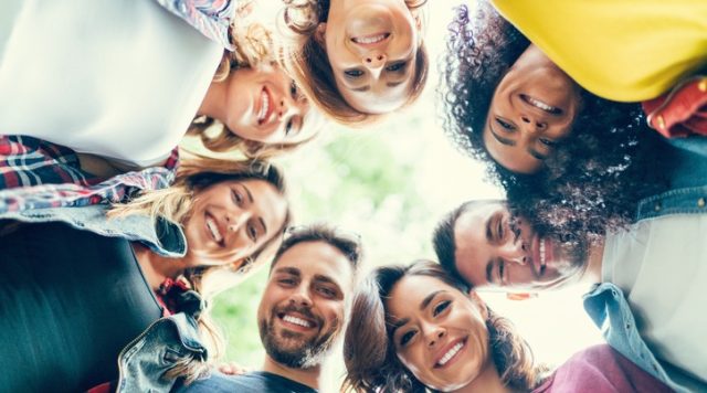 A group of friends standing in a circle with their heads together, looking down and smiling at the camera