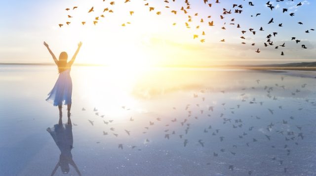 Photo of a happy woman with her hands up in the air. She is standing on the beach with the sun shining brightly and birds flying in the sky.