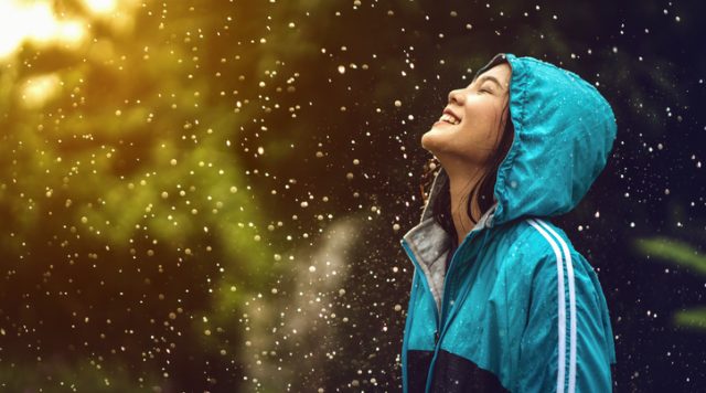 Photo of a young woman in a raincoat standing outside in a light rain while the sun shines through. Her head is tilted up to the sky and she is smiling