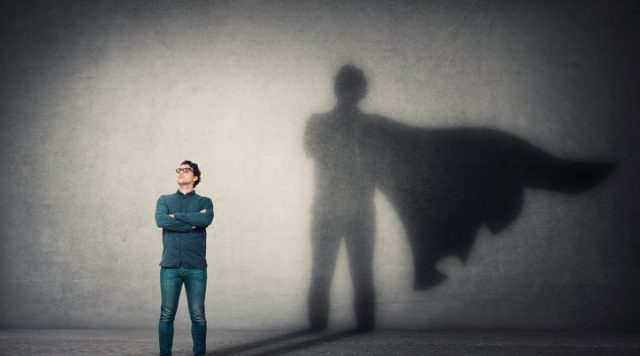Photo of a man standing with his arms crossed, his shadow cast on a wall behind him has the addition of a superhero cape