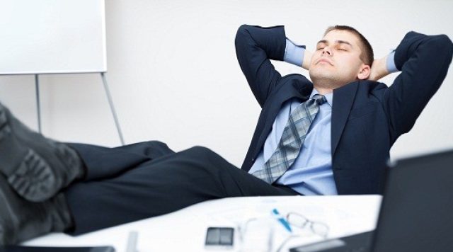 A man in a suit sitting at a desk, leaning back with his feet up and his arms behind his head
