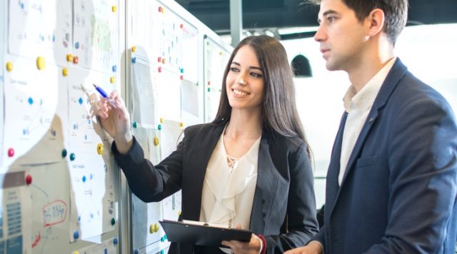A man and a woman, both in business suits, standing looking at a whiteboard. The woman is teaching the man something