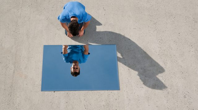 A mirror on the ground with a man crouching down looking into it at his reflection