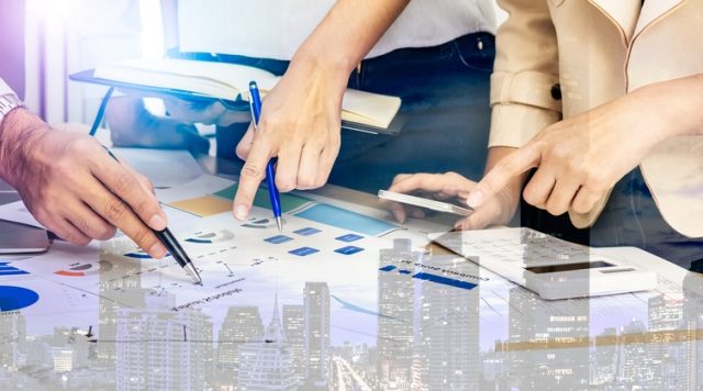 Photo of 3 people standing around a table pointing at charts and data