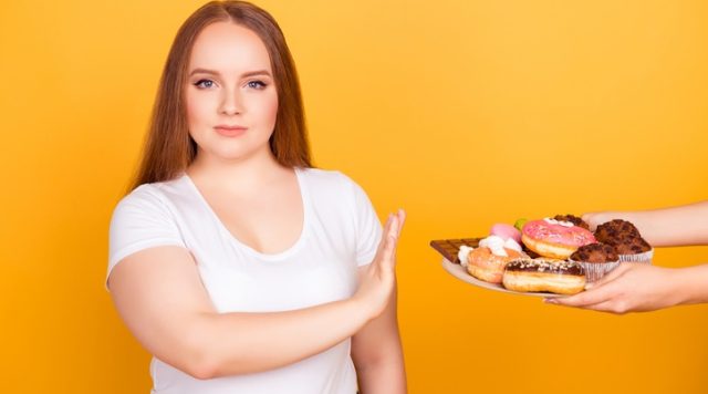 I'm against eating products containing fat! Will-powered woman wearing white tshirt is refusing to consume tasty delicious sweets on a plate, isolated on bright yellow background
